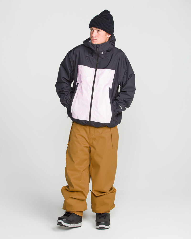 A person wearing a black beanie, a black and white recycled twill jacket, Volcom Men’s Arthur 20K Pants in brown, and black sneakers stands against a plain white background with their hands in their jacket pockets.