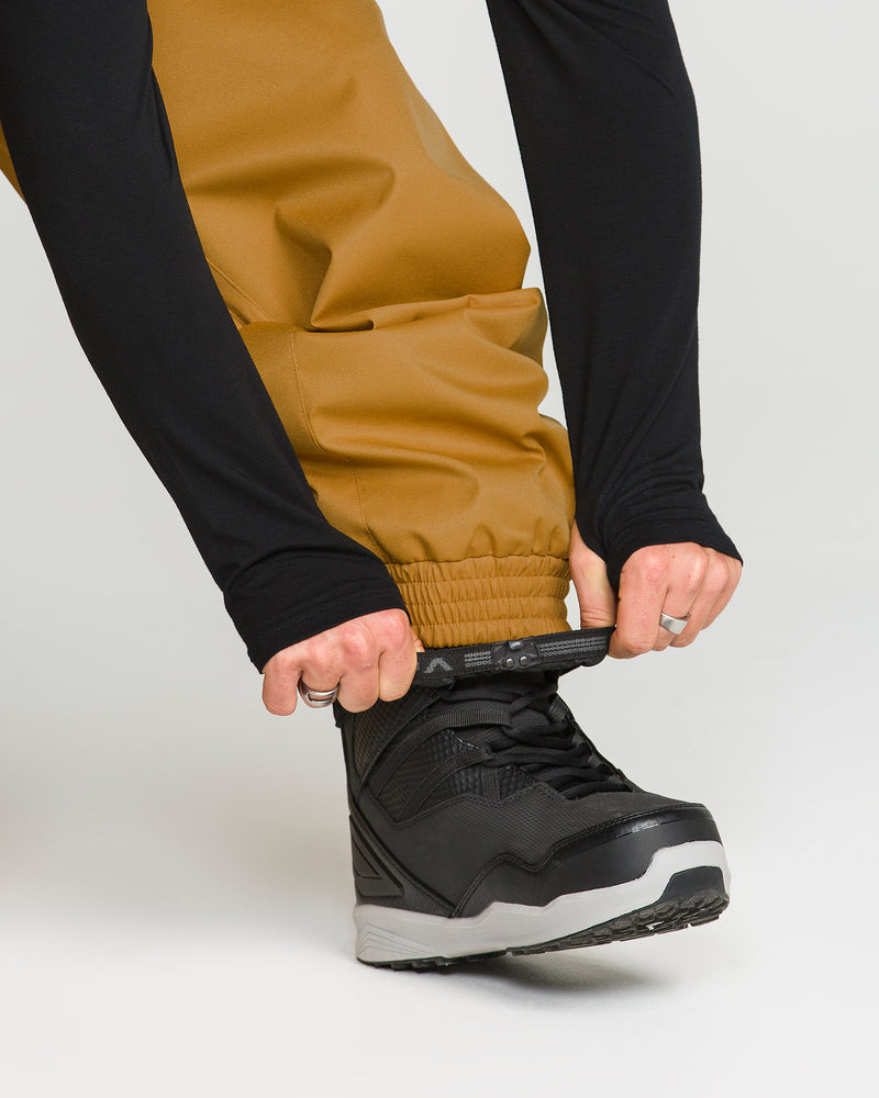 A person wearing tan Volcom Men’s Arthur 20K Pants made from recycled twill and black long sleeves adjusts the elastic cuff over a black and gray sneaker-style boot.
