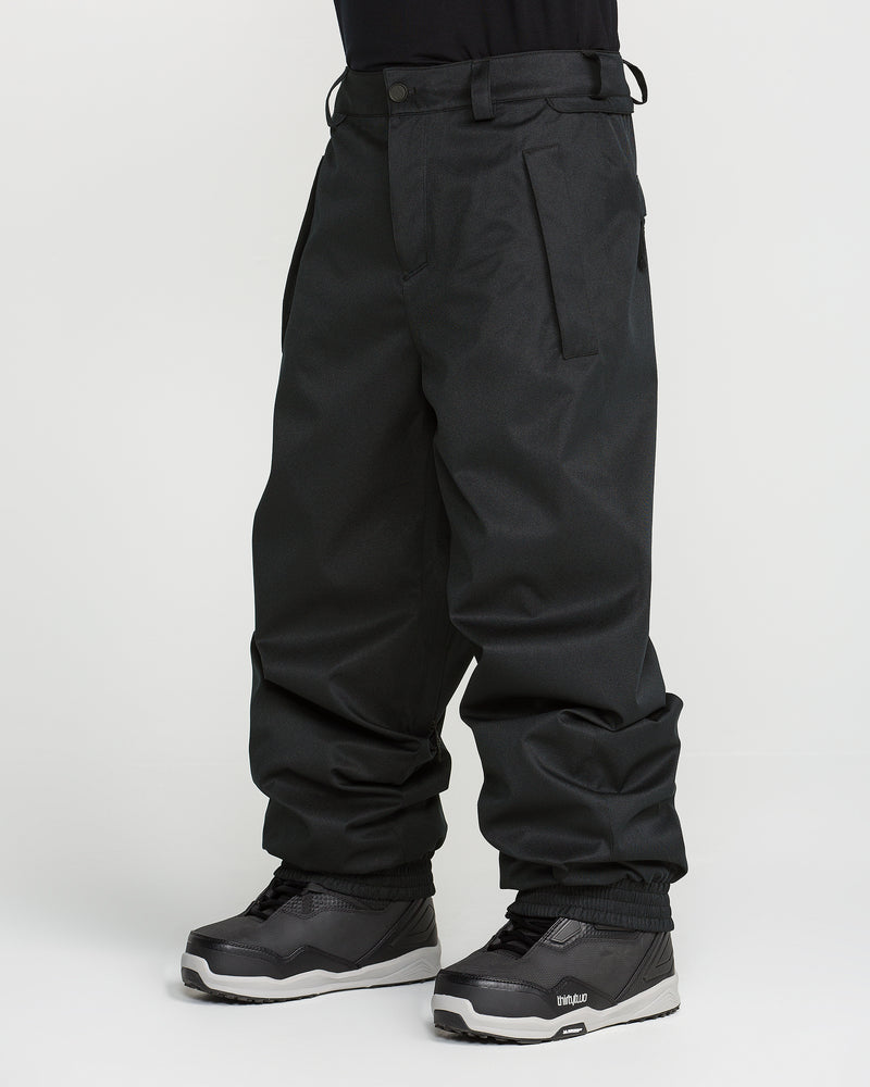A person wearing Volcom Men’s Arthur 20K Pants in loose-fitting black recycled twill and black snowboard boots, standing against a plain white background.