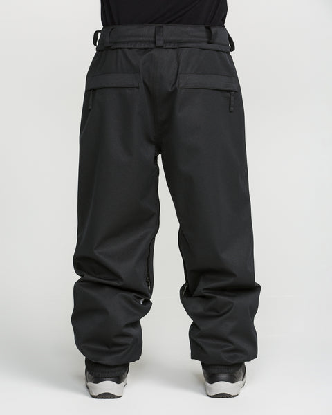 A person wearing loose-fitting black Volcom Men’s Arthur 20K Pants, featuring zipper pockets and crafted from recycled twill, paired with white sneakers and photographed from the waist down against a plain white background.