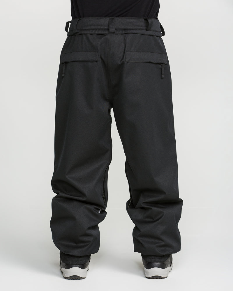 A person wearing loose-fitting black Volcom Men’s Arthur 20K Pants, featuring zipper pockets and crafted from recycled twill, paired with white sneakers and photographed from the waist down against a plain white background.