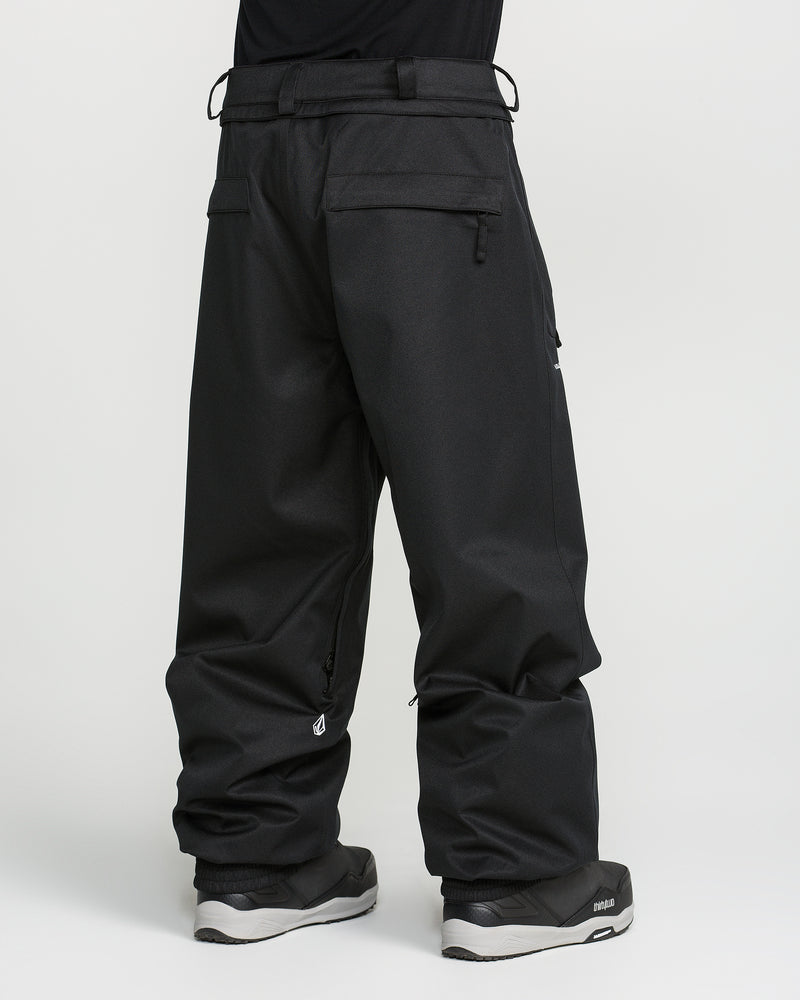 A person is seen from behind wearing loose-fitting black Volcom Men’s Arthur 20K Pants, crafted from recycled twill, paired with black sneakers against a plain white background. The pants feature zippered pockets and belt loops, and their hems drape over most of the shoes.