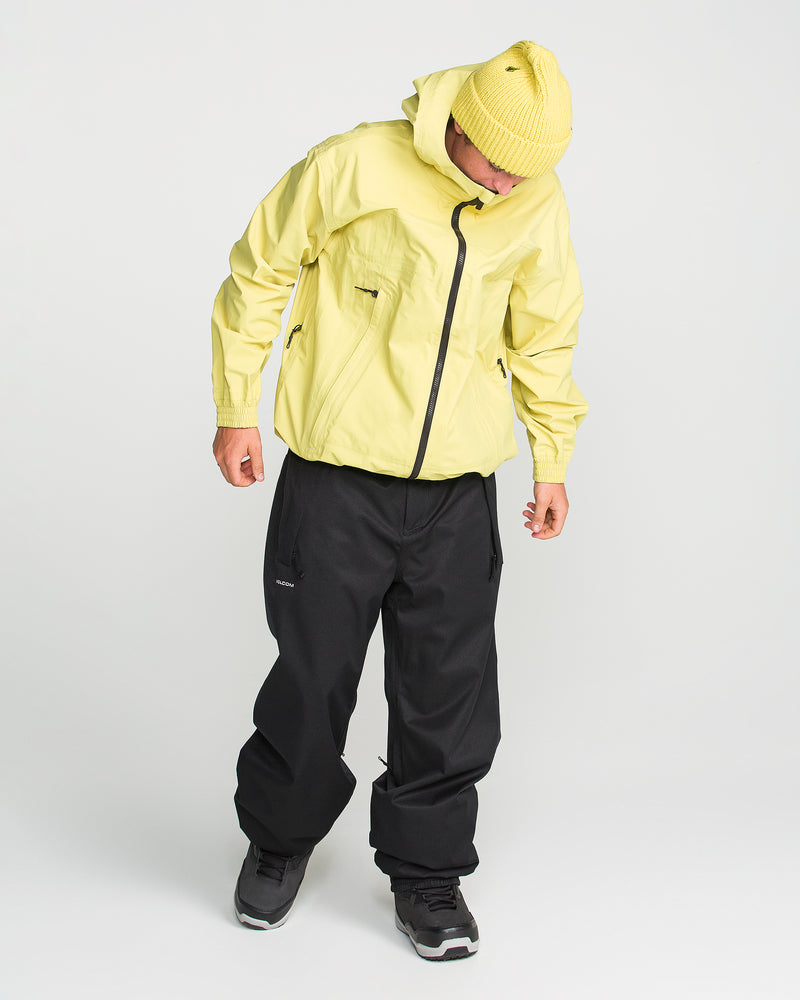 Wearing a yellow hooded jacket, yellow beanie, black Volcom Men’s Arthur 20K Pants made from recycled twill, and black sneakers, a person stands looking down against a plain white background.