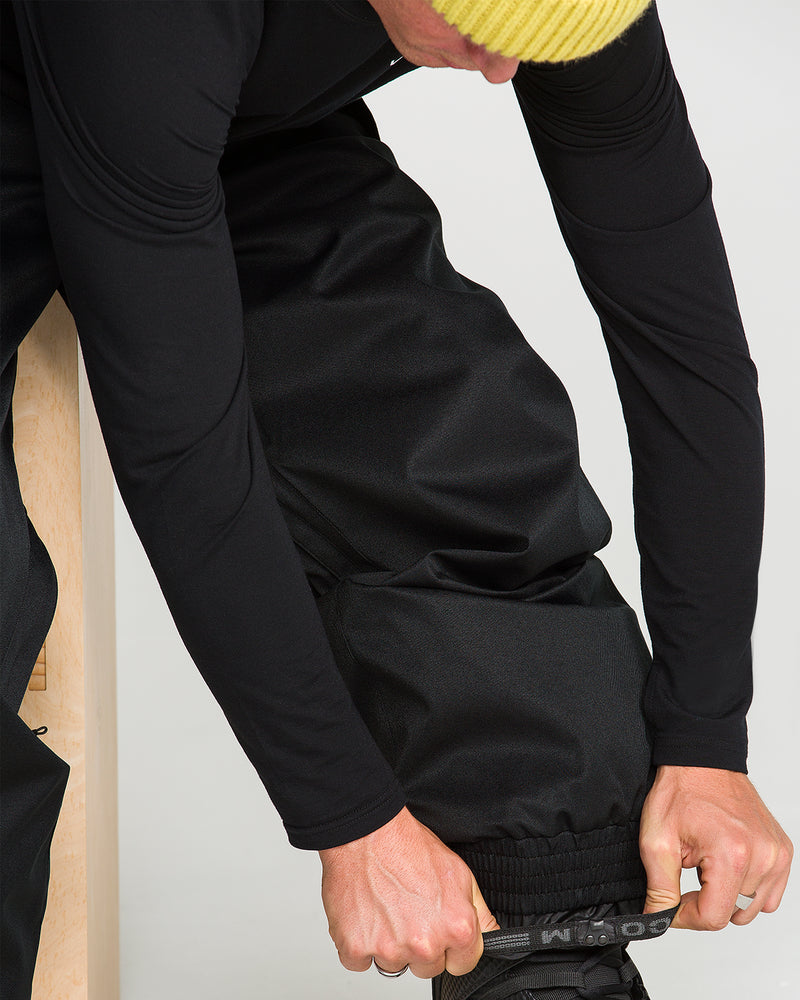 A person wearing a yellow knit hat and black long-sleeve clothes bends down to adjust the hem of their black Volcom Men’s Arthur 20K Pants over a boot.