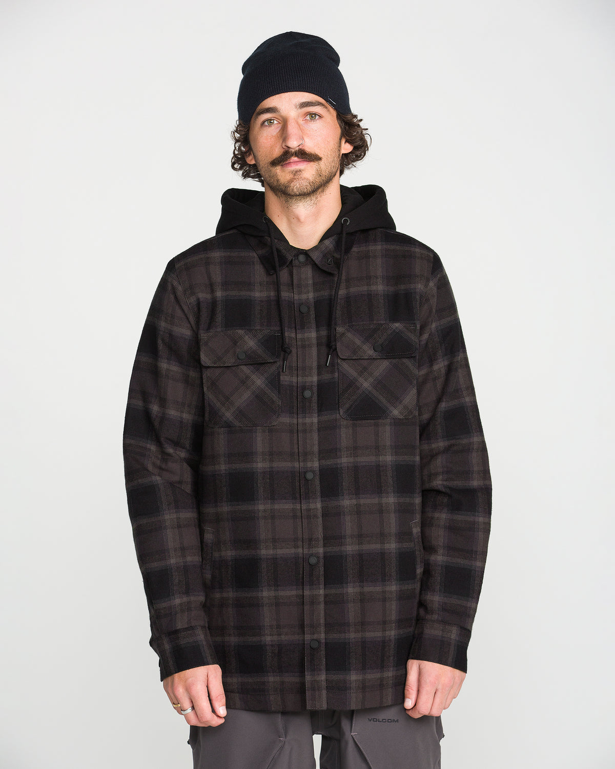 A man with a mustache, wearing a black beanie, Volcom Men’s Enson Insulated Riding Flannel in a black and gray plaid pattern, and gray pants stands facing forward against a plain white background.