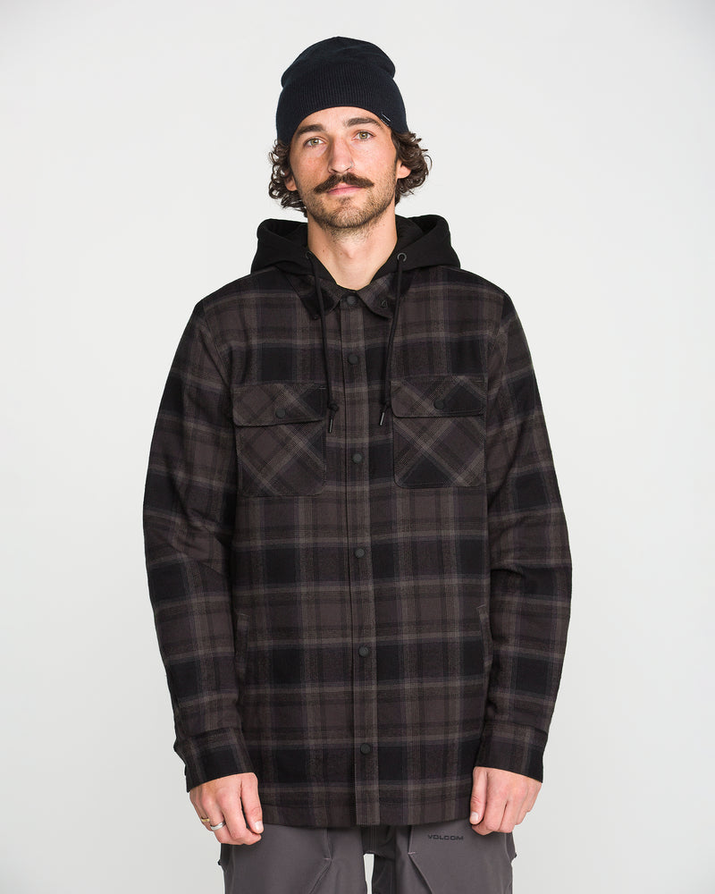 A man with a mustache, wearing a black beanie, Volcom Men’s Enson Insulated Riding Flannel in a black and gray plaid pattern, and gray pants stands facing forward against a plain white background.