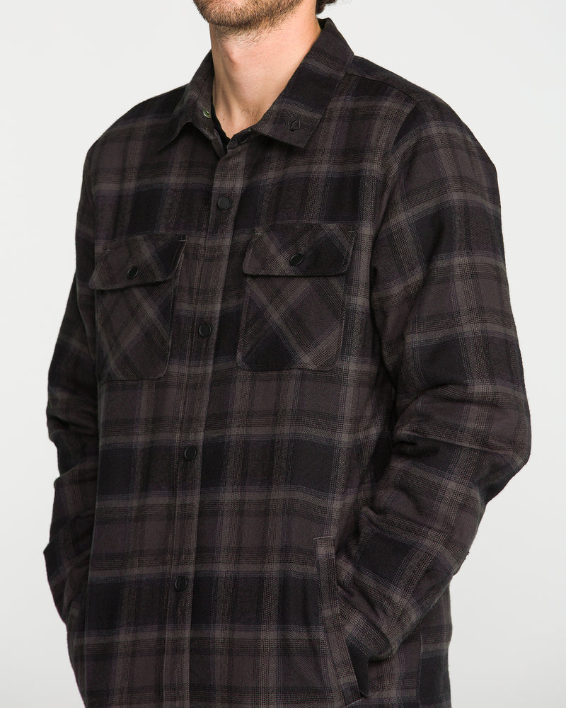 A man wearing the Volcom Men’s Enson Insulated Riding Flannel, a dark plaid button-up shirt with flap chest pockets, stands with his hands in his pockets. His face is partially out of frame.