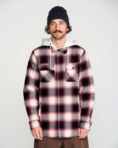 A man stands against a plain light background wearing a black beanie, a white hoodie with a cozy fleece hood, and the Volcom Men’s Enson Insulated Riding Flannel—a red, black, and white button-up shirt with two chest pockets.