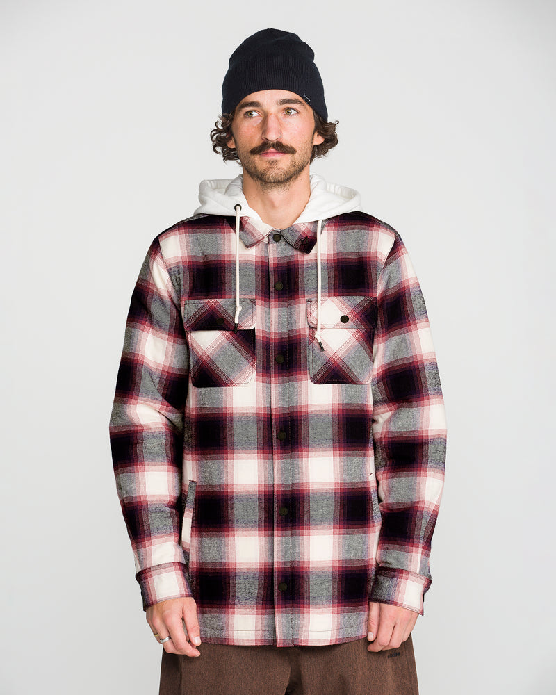 A man stands against a plain light background wearing a black beanie, a white hoodie with a cozy fleece hood, and the Volcom Men’s Enson Insulated Riding Flannel—a red, black, and white button-up shirt with two chest pockets.