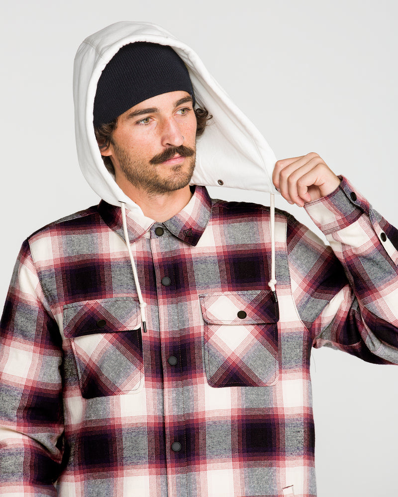 A man with a mustache wears a black beanie and the Volcom Men’s Enson Insulated Riding Flannel, featuring a plaid pattern and white fleece hood, holding up the hood with one hand against a plain background.