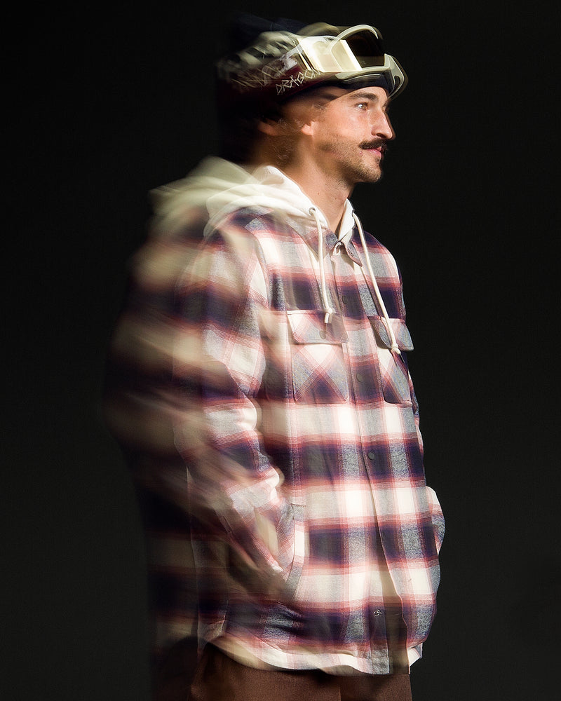 A man stands against a dark background, wearing the Volcom Men’s Enson Insulated Riding Flannel over a white hoodie with a fleece hood and ski goggles. A blur effect gives his left side a sense of motion.