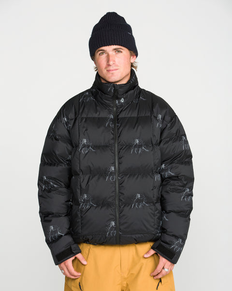 A person stands against a plain white background with hands in pockets, looking at the camera, wearing a black beanie, tan pants, and the Volcom Men’s Arthur Down Puff Jacket—a PFAS-free DWR black puffer jacket with subtle patterns.