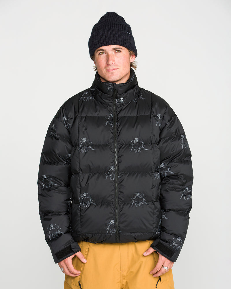 A person stands against a plain white background with hands in pockets, looking at the camera, wearing a black beanie, tan pants, and the Volcom Men’s Arthur Down Puff Jacket—a PFAS-free DWR black puffer jacket with subtle patterns.