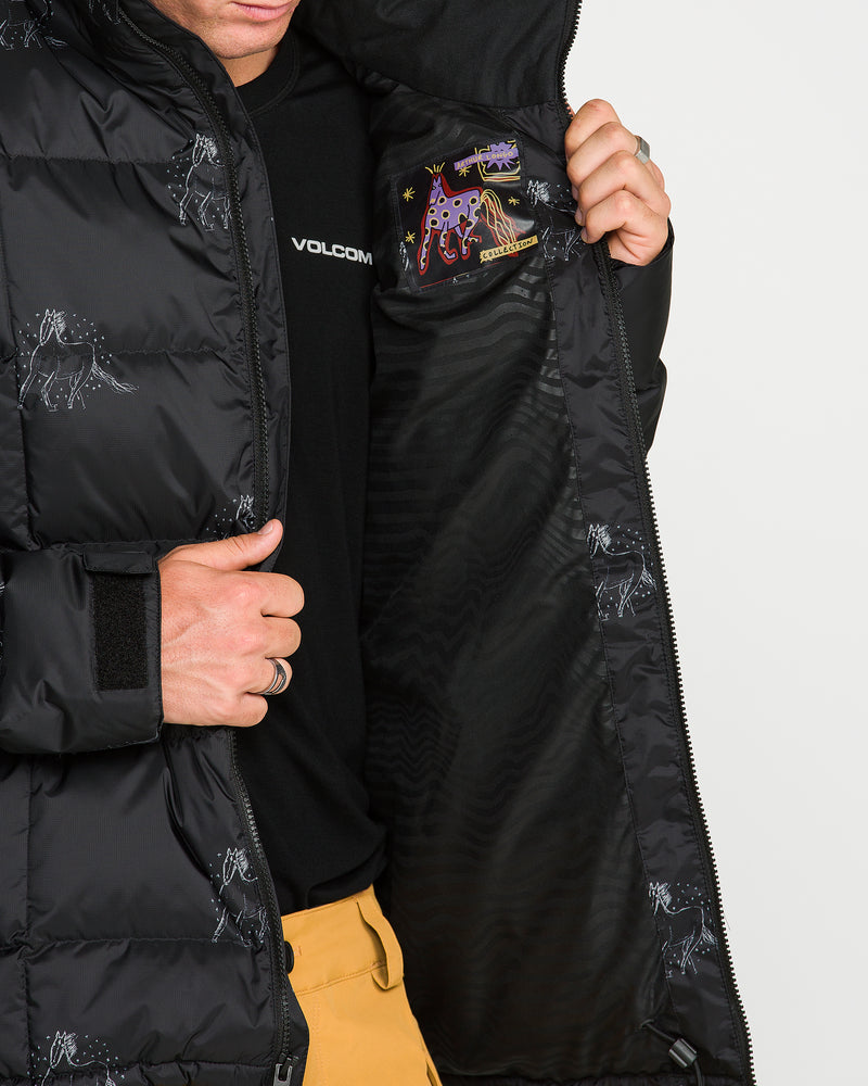 Wearing Volcom’s Men’s Arthur Down Puff Jacket, a person holds the jacket open to reveal its constellation-themed patch on the inner lining. The eco-friendly jacket features PFAS-free DWR for water resistance and is paired with tan pants, while the individual’s face is partially out of frame.