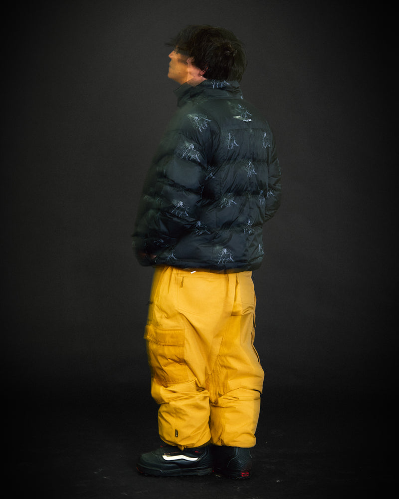 Against a dark background, a person wears Volcom’s Men’s Arthur Down Puff Jacket in black, paired with loose-fitting yellow pants. Their relaxed pose and slightly turned head evoke the signature style of Arthur Longo.
