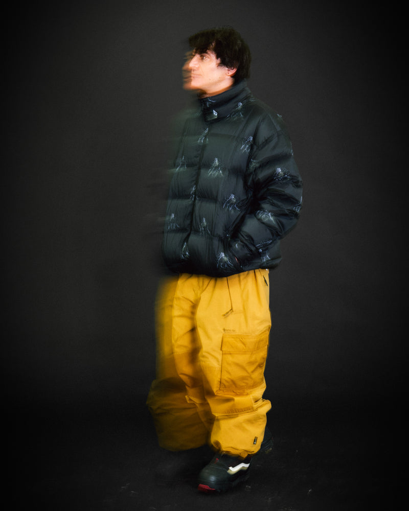 Wearing the Volcom Men’s Arthur Down Puff Jacket in a V-Science 2-Layer dark puffer style paired with baggy yellow pants, a person stands against a dark background. Their blurred face and upper body suggest motion, capturing the signature baggy-fit down puff look inspired by Arthur Longo.