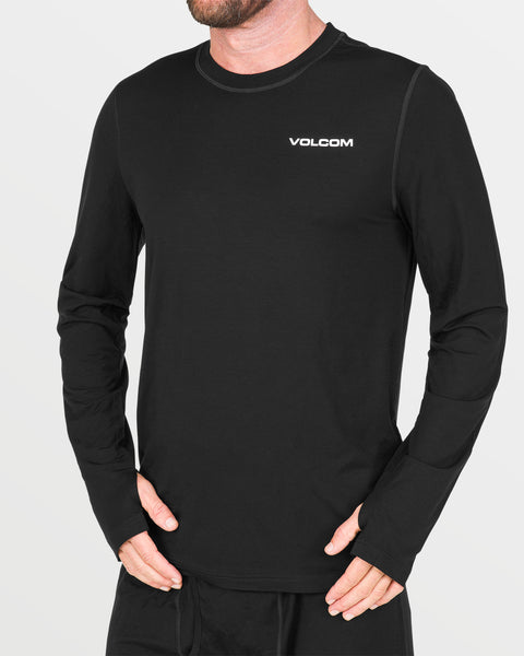 A person is wearing the Volcom Men’s M Merino Blend Crew, a black long-sleeve base layer featuring “VOLCOM” in white on the chest. Designed for men, this shirt includes thumbholes for enhanced comfort and practicality. The individual is standing against a plain light background.