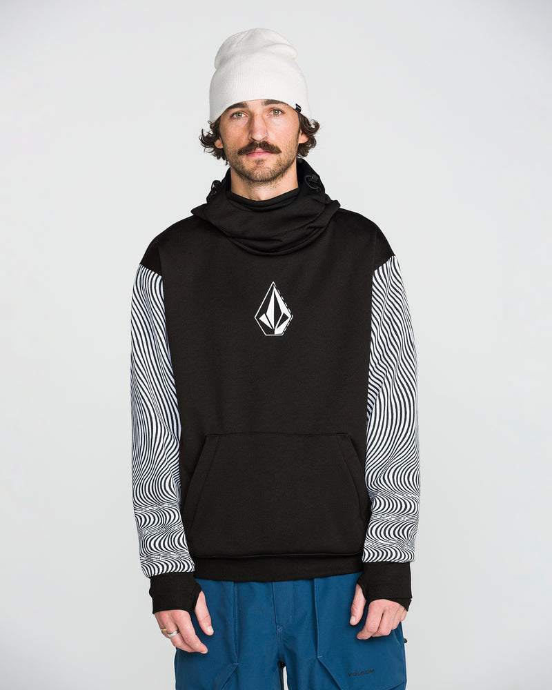 A man wearing the Volcom Men’s Hydro Riding Hoodie featuring a geometric Volcom Stone logo and patterned sleeves, paired with a white beanie and blue pants, stands against a plain light background.