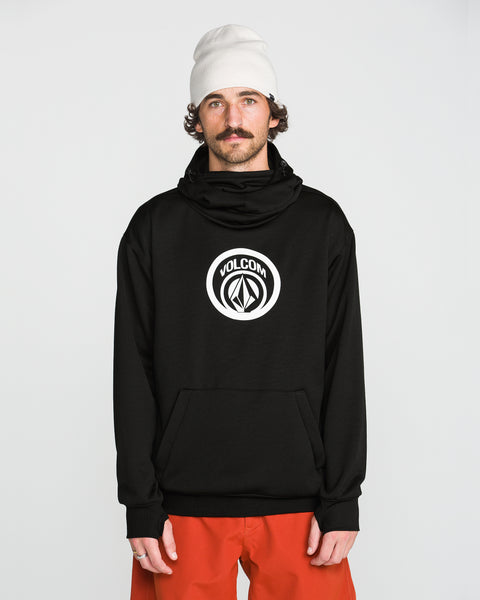A man wearing a white beanie, orange pants, and a black Volcom Men’s Hydro Riding Hoodie with a white logo stands facing forward against a plain white background.