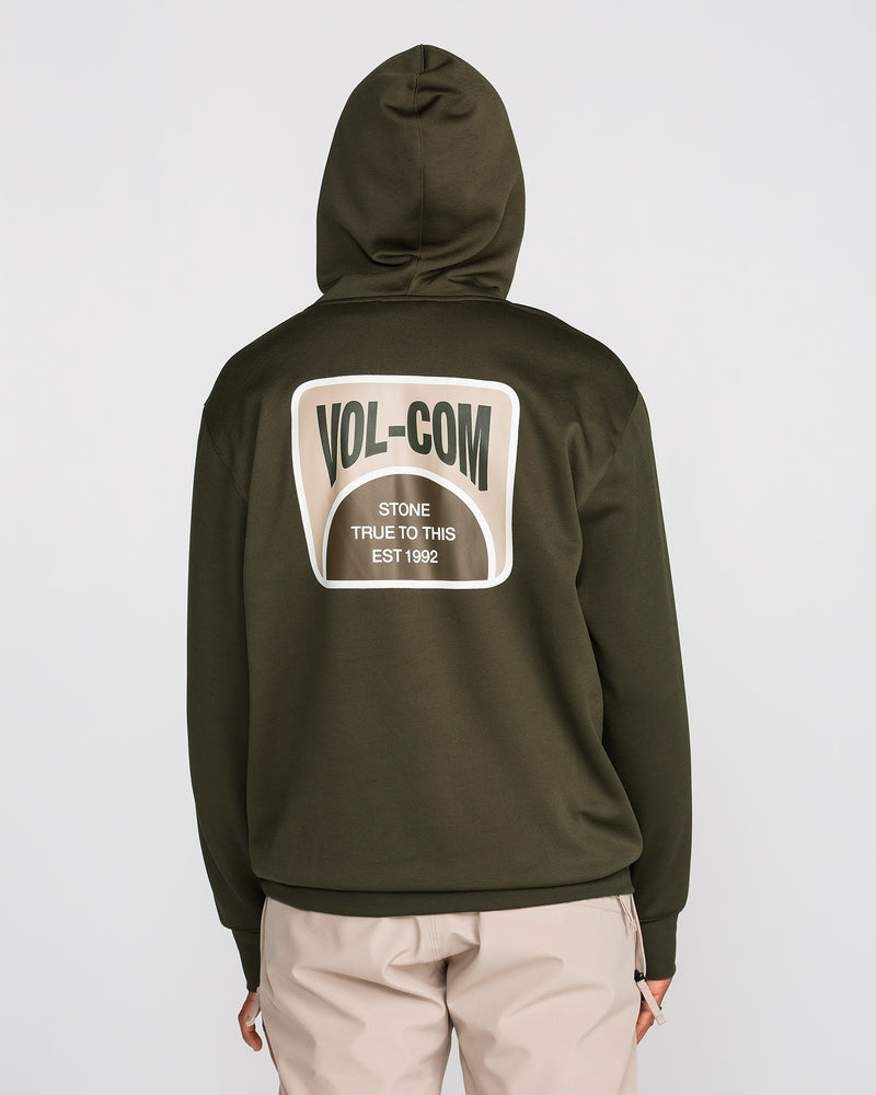 A person is wearing a Volcom Men’s Hydro Riding Hoodie in olive green with the hood up. The back of the hoodie features a large patch that reads “VOL-COM Stone True To This EST 1992.” They are also wearing beige pants.