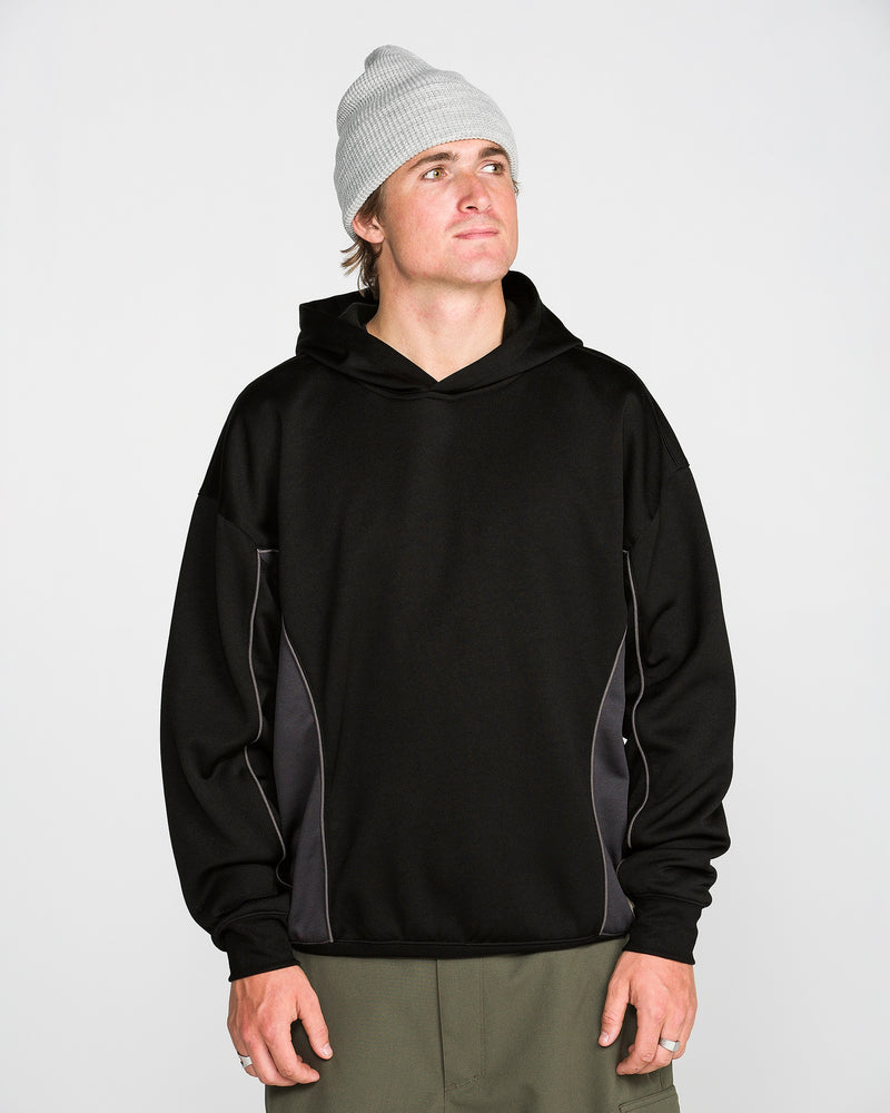 A person wearing a light gray beanie, olive green pants, and the Volcom Men’s Vital Hydro Pullover Hoodie in black with gray side panels stands against a plain white background, looking slightly upward and to the right.