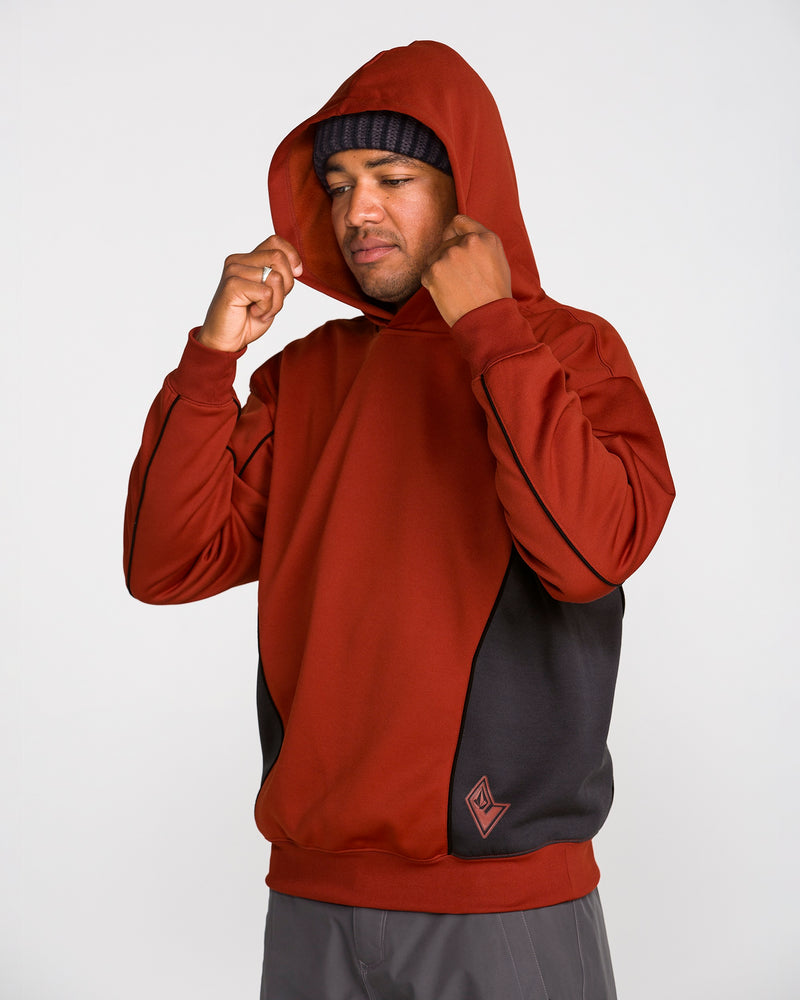 A person wearing the Volcom Men’s Vital Hydro Pullover in red and black, along with a black beanie, adjusts their hood while standing against a plain, light-colored background.