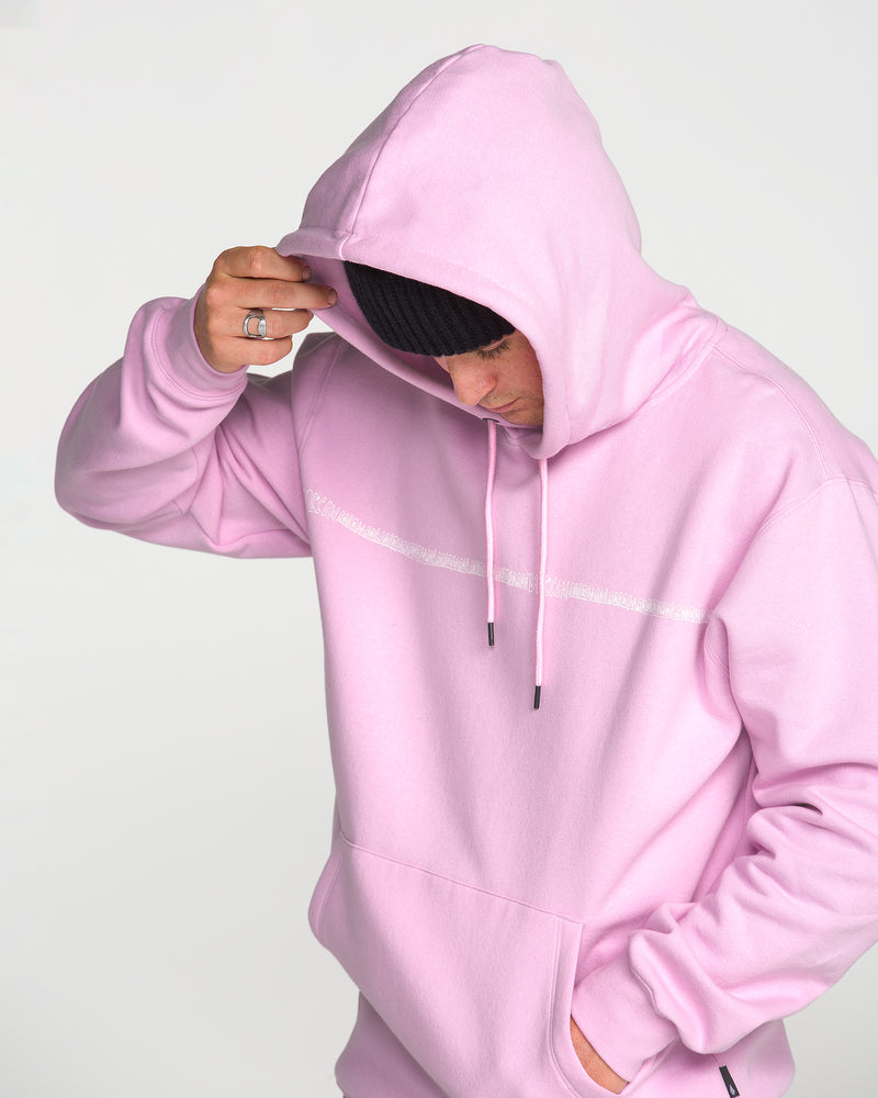 A person wearing the Volcom Men’s Essential Hoodie in light pink, crafted from soft cotton-poly fleece, along with a black beanie, tilts their head down and holds the hood with one hand against a plain light background.