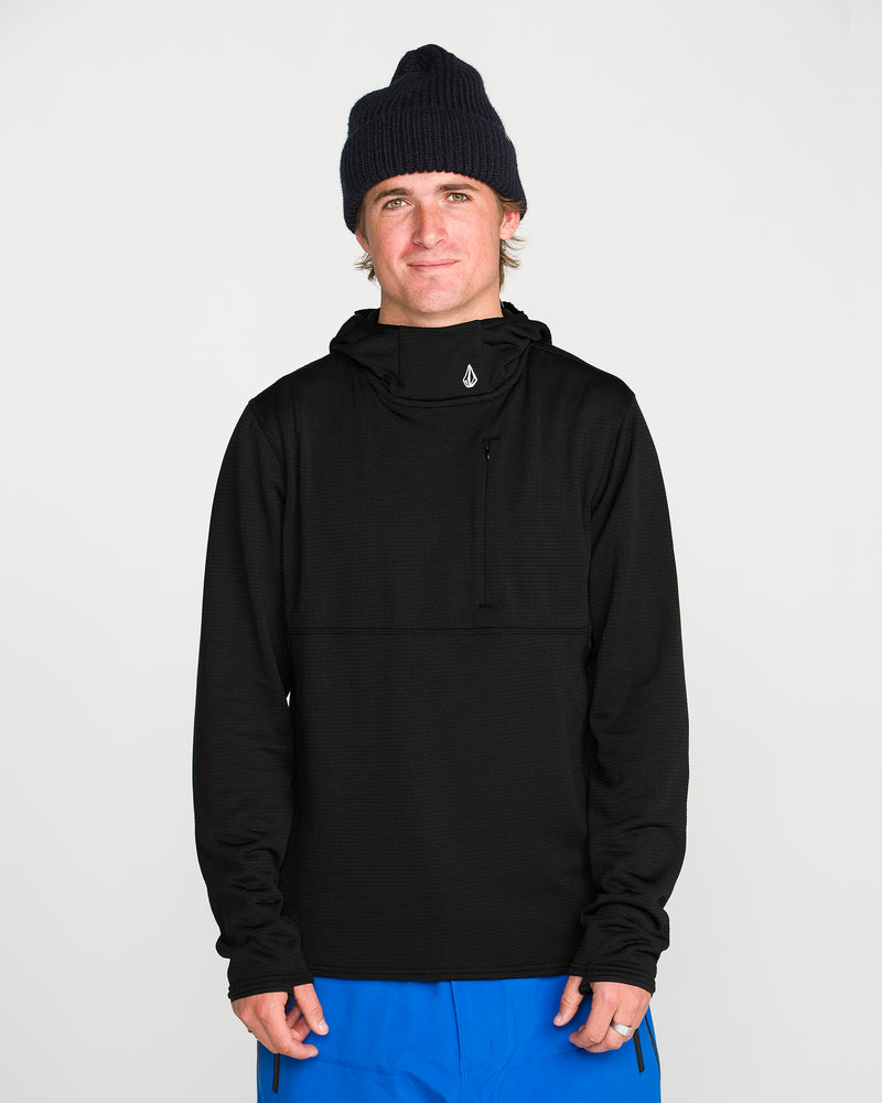 A person wearing a black Volcom Men’s Gridlock Hooded Pullover, a black knit beanie, and blue pants stands against a plain white background, facing forward with a neutral expression.