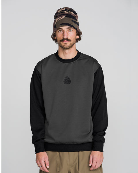 A man wearing the Volcom Men’s Hydro Crew, a dark gray and black sweatshirt made from hydrophobic fleece for moisture-wicking comfort and dryness all day, pairs it with beige pants and a brown patterned beanie while standing against a plain light gray background and looking at the camera with a neutral expression.