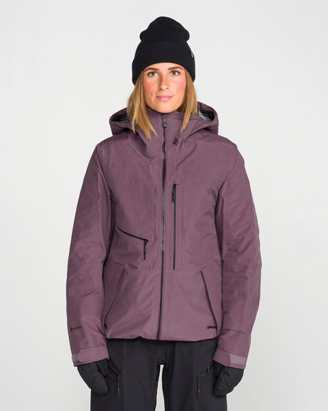 A person is wearing the Volcom Women’s Koa TDS Infrared Gore-Tex Jacket in dark purple, paired with a black beanie and black gloves, standing facing forward against a plain light background.