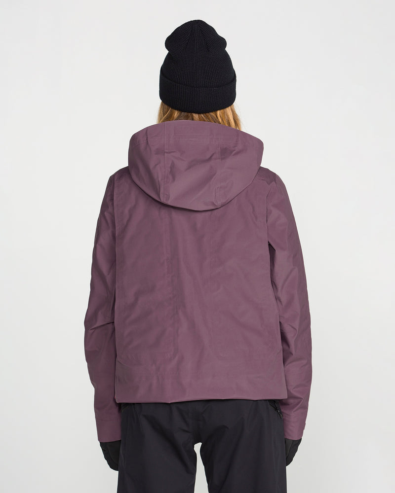 A person wearing the Volcom Women’s Koa TDS Infrared Gore-Tex Jacket in purple, paired with black pants, black gloves, and a black beanie, stands with their back to the camera against a plain white background.