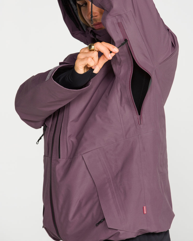 A person wearing the Volcom Women’s Koa TDS Infrared Gore-Tex Jacket in purple uses their right hand to unzip an underarm vent on the left side. The image highlights the jacket’s detailed features, such as zippers and pockets, and showcases its advanced Thermal Defense System for enhanced comfort and protection.