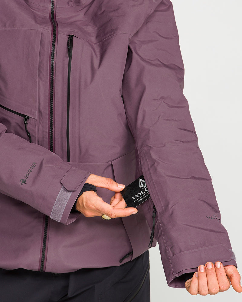 A woman wearing the Volcom Women’s Koa TDS Infrared GORE-TEX Jacket, featuring a thermal defense system, reveals a black Volcom tag inside the sleeve pocket by pulling it open with one hand while extending her other arm.