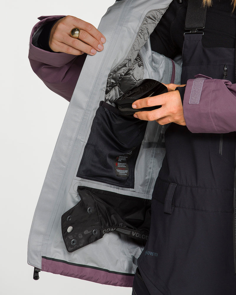 A person opens a Volcom Women’s Koa TDS Infrared Gore-Tex Jacket in purple and black, revealing an inner pocket with a black pouch inside. The light gray lining, featuring a silver section, hints at the advanced thermal defense system integral to this GORE-TEX jacket. Only the person’s hands and part of their torso are visible.