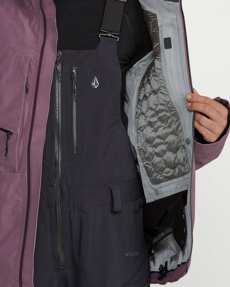 A person wearing dark winter overalls and the Volcom Women’s Koa TDS Infrared Gore-Tex Jacket in purple holds the jacket open to reveal its gray quilted inner lining and an interior pocket with black gloves, showcasing the jacket’s advanced thermal defense system.