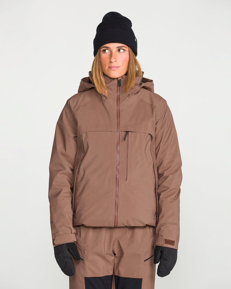 A person is wearing the Volcom Women’s 3D Stretch Gore Jacket in brown, paired with matching pants, black gloves, and a black knit beanie. They are standing against a plain white background and looking slightly to the side.