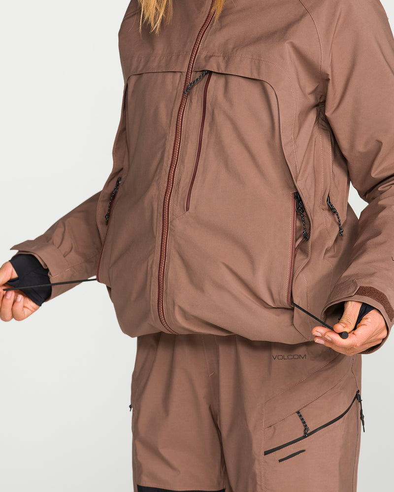 A person wearing the Volcom Women’s 3D Stretch Gore Jacket in brown is adjusting the drawstrings at the bottom of the jacket. Only their torso and hands are visible. The GORE-TEX jacket features zipped pockets, and the matching pants have zippered details.