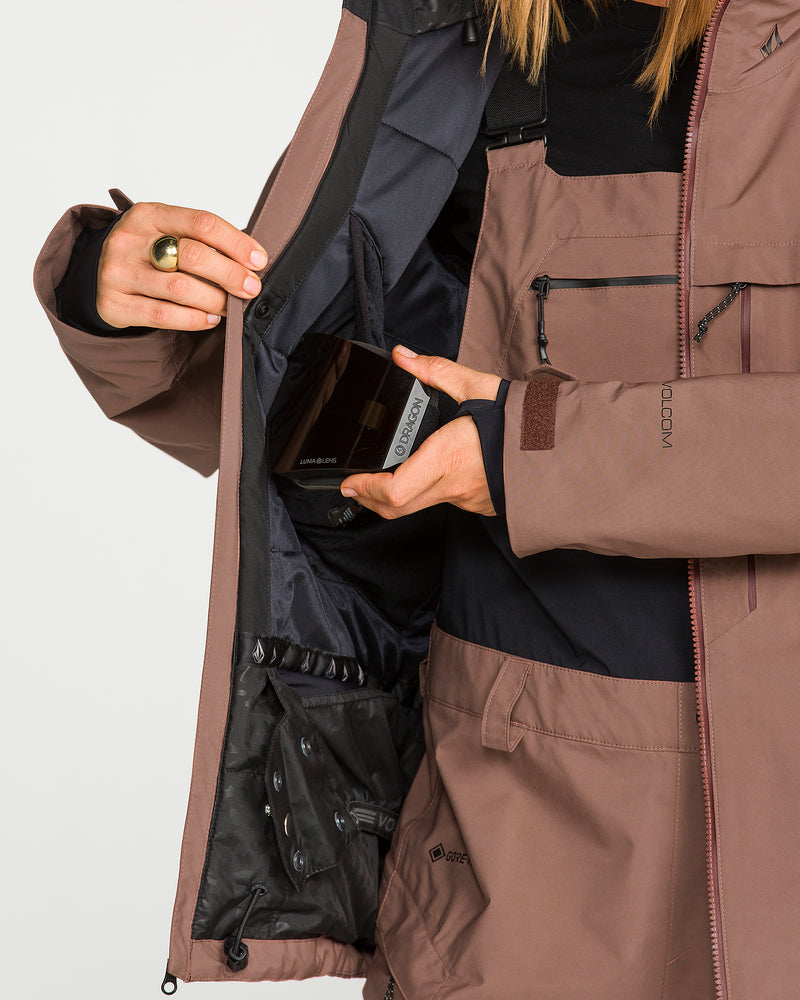 A person wearing the Volcom Women’s 3D Stretch Gore Jacket in brown opens an inner pocket to place a rectangular item inside, revealing the jacket’s mesh pouch and black inner lining. Only the torso and hands are visible.