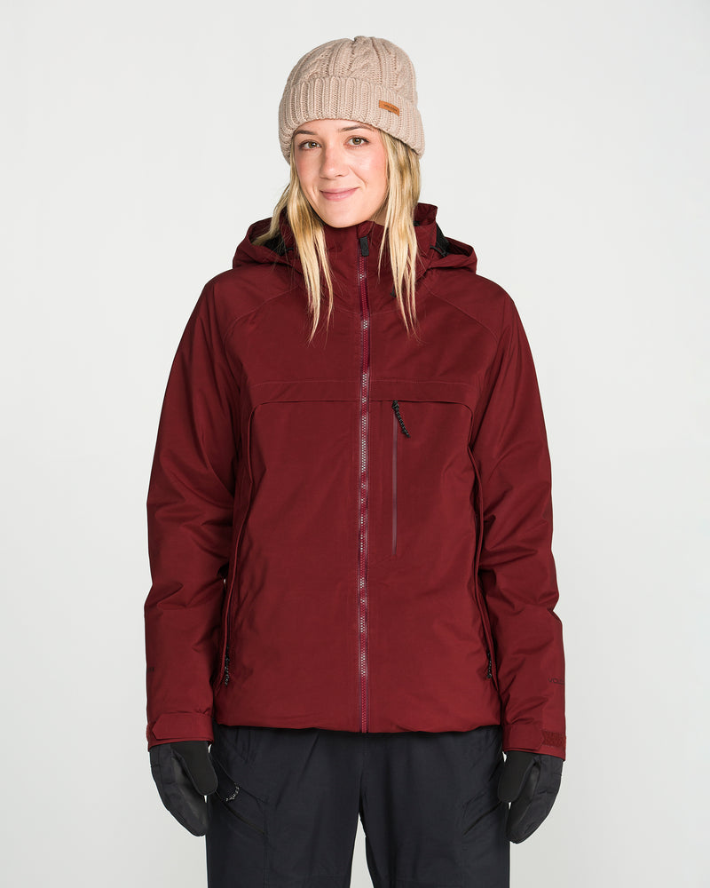 A person wearing the Volcom Women’s 3D Stretch Gore Jacket in maroon, paired with black gloves and a beige knit beanie, stands against a plain white background, smiling softly.