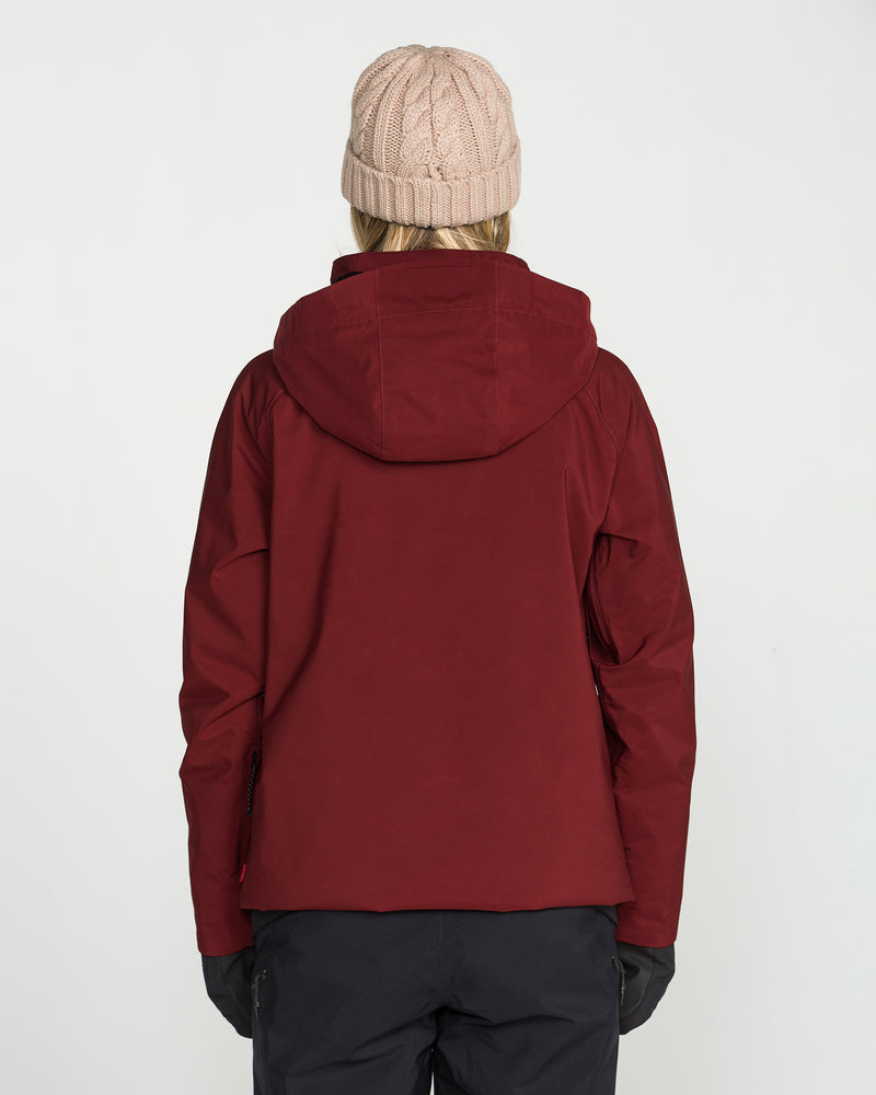 A person is seen from behind against a plain white background, wearing the Volcom Women’s 3D Stretch Gore Jacket in maroon, paired with black pants, black gloves, and a beige knit beanie.