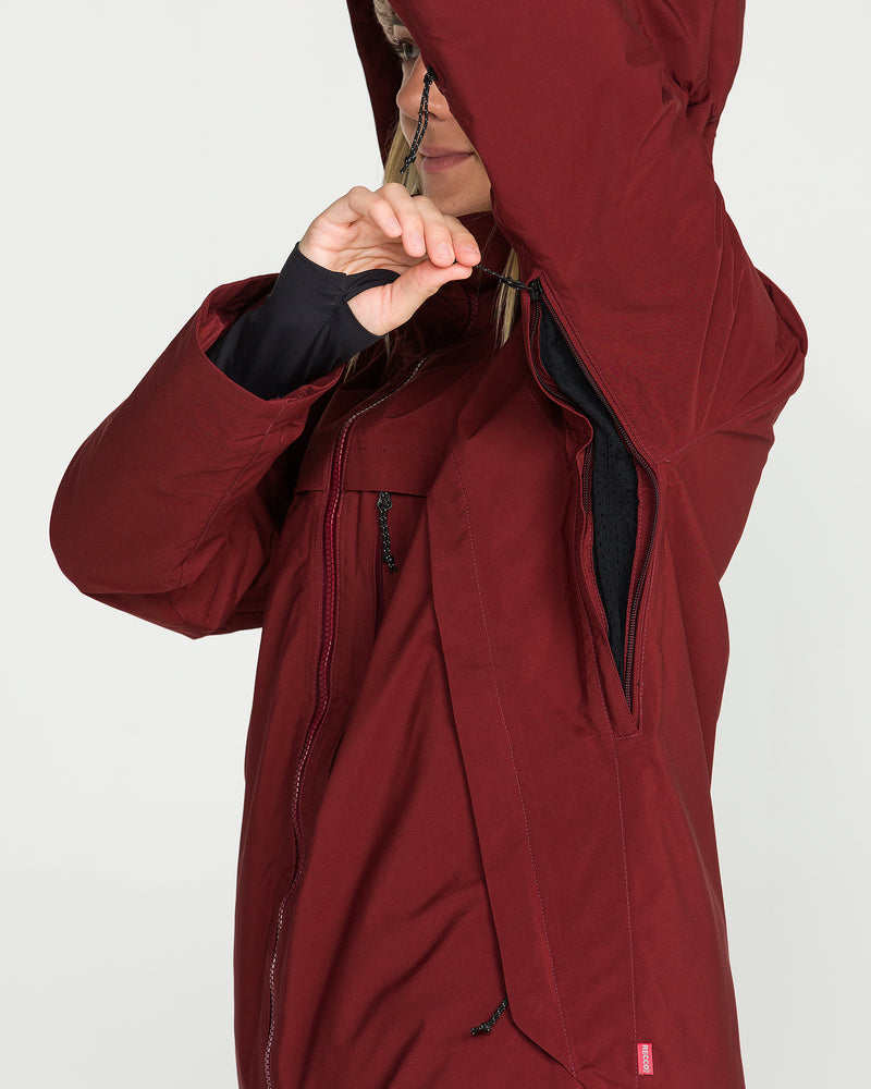 A person wearing the Volcom Women’s 3D Stretch Gore Jacket in maroon demonstrates the armpit zipper ventilation with the hood partially raised; only their hands and the lower part of their face are visible.