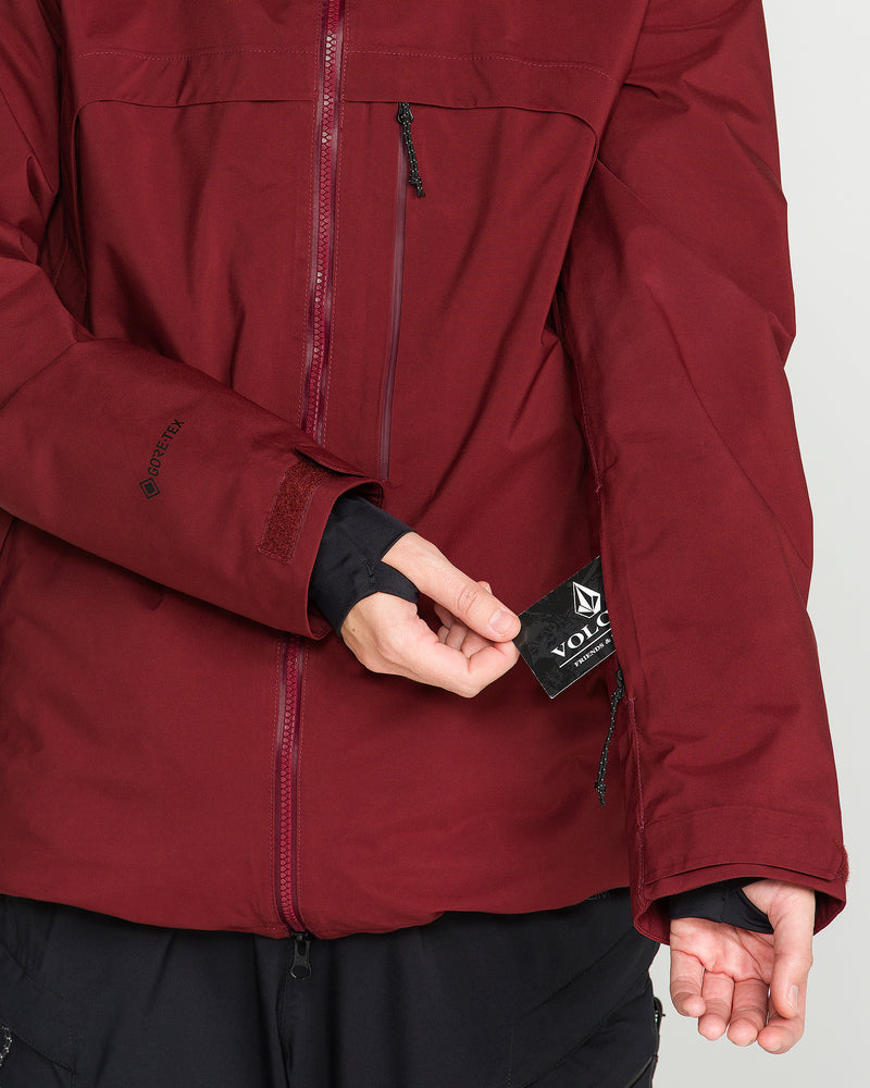 A person wearing the Volcom Women’s 3D Stretch Gore Jacket in maroon is pulling out a small black Volcom tag from a zippered pocket on the sleeve.
