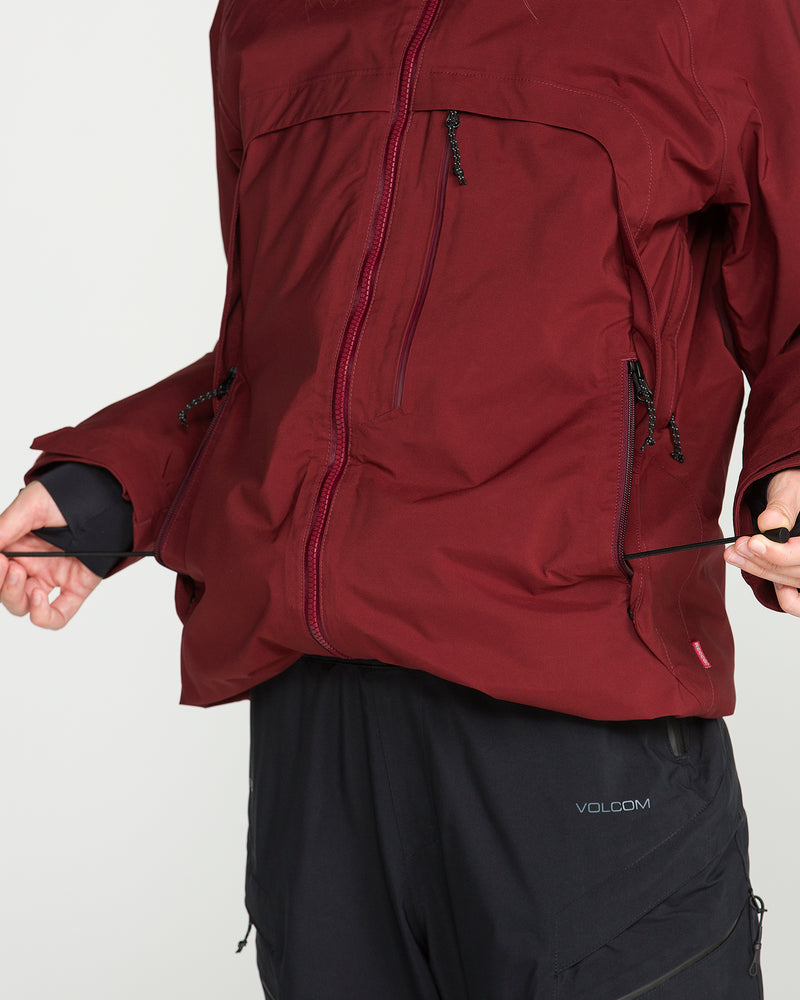 A person adjusts the drawstring on a maroon Volcom Women’s 3D Stretch Gore Jacket featuring waterproof construction, zippered pockets, and PrimaLoft insulation, paired with black pants. Only the torso and hands are visible against a plain background.