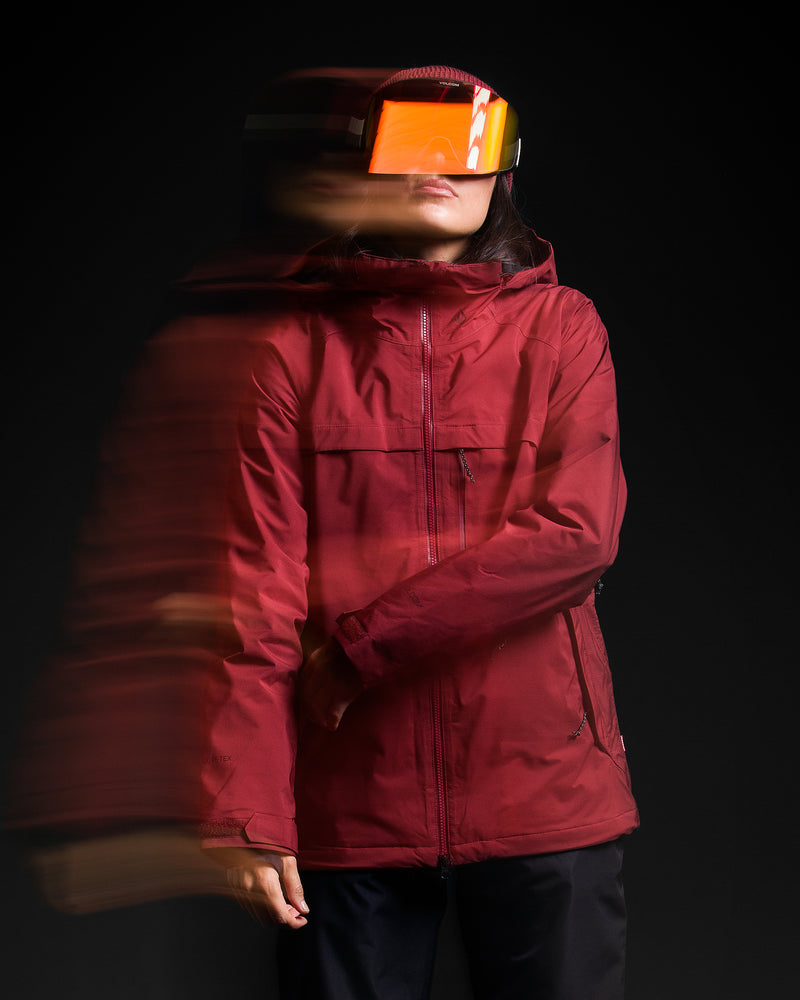 Wearing the Volcom Women’s 3D Stretch Gore-Tex Jacket in red and a large, reflective orange visor, a person stands against a black background with a motion blur effect on their left side.