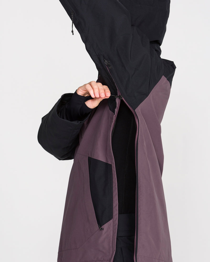 A person wearing the Volcom Women’s Rowan Insulated Gore Pullover in purple and black zips up the front while raising the hood over their head, partially covering their face.