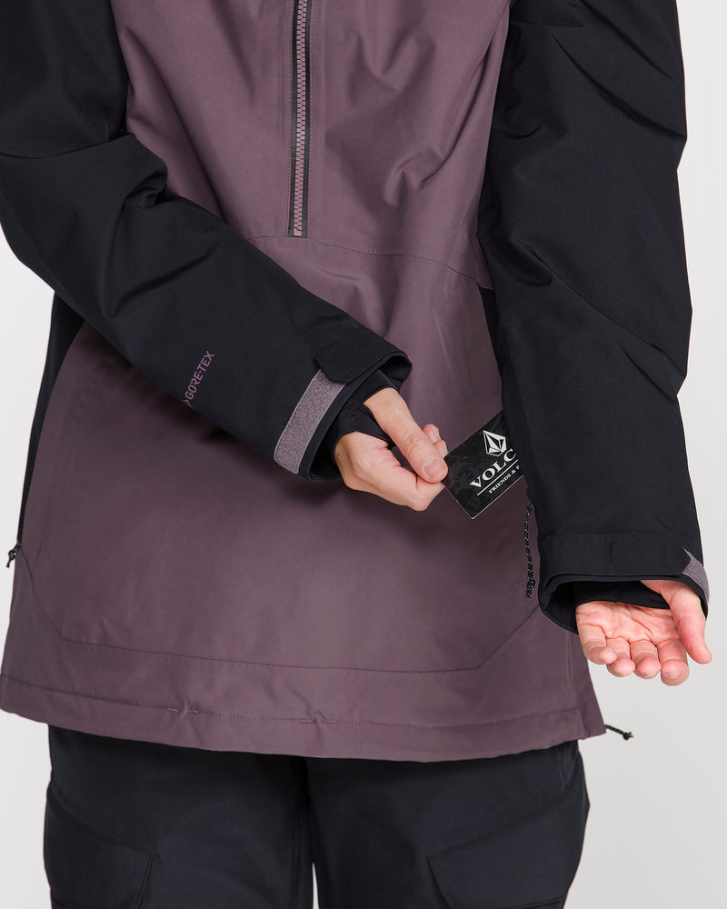 A person wearing the Volcom Women’s Rowan Insulated Gore Pullover in purple and black pulls on a Volcom logo tag attached to the jacket’s sleeve, with their other palm facing up.