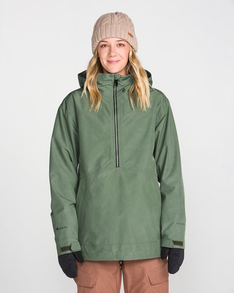 A woman stands facing forward against a plain white background, smiling slightly while wearing the Volcom Women’s Rowan Insulated Gore Pullover in green, paired with a beige knit beanie, brown pants, and black gloves.