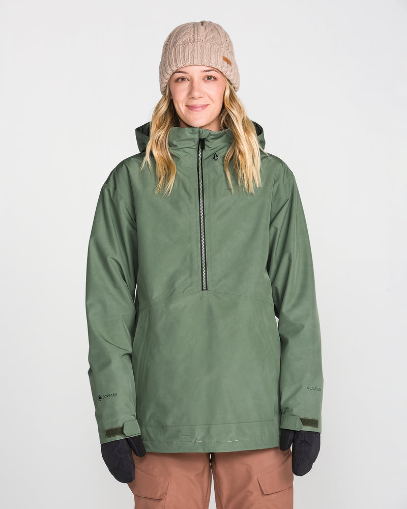 A woman stands facing forward against a plain white background, smiling slightly while wearing the Volcom Women’s Rowan Insulated Gore Pullover in green, paired with a beige knit beanie, brown pants, and black gloves.