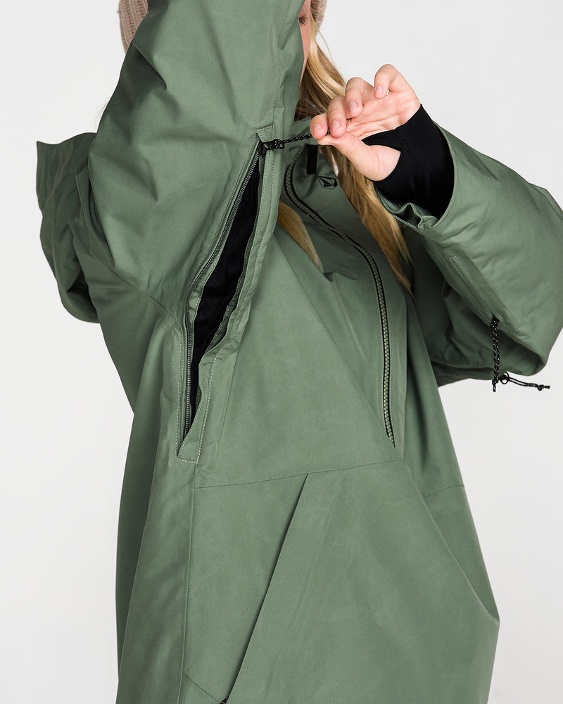 A person wearing the Volcom Women’s Rowan Insulated Gore Pullover in green unzips an underarm vent. Their face is partially obscured by a raised arm and the jacket’s hood, set against a plain white background.