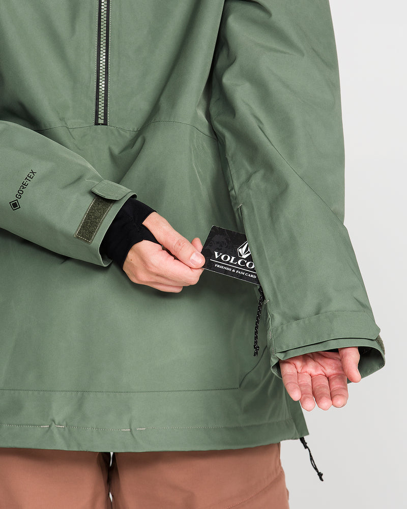 Wearing the Volcom Women’s Rowan Insulated Gore Pullover, a person in a green waterproof ski jacket displays a Volcom tag from an inner pocket, highlighting the jacket’s features, including its visible zipper and Velcro strap on the sleeve.
